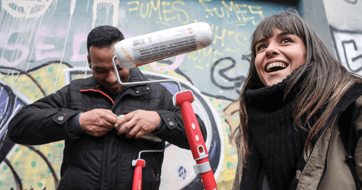 Imagen que muestra una acción de intervención comunitaria llevada a cabo por Emaús. En la fotografia se ve una chica y un chico con un rodete de pintura delante de un muro con grafittis